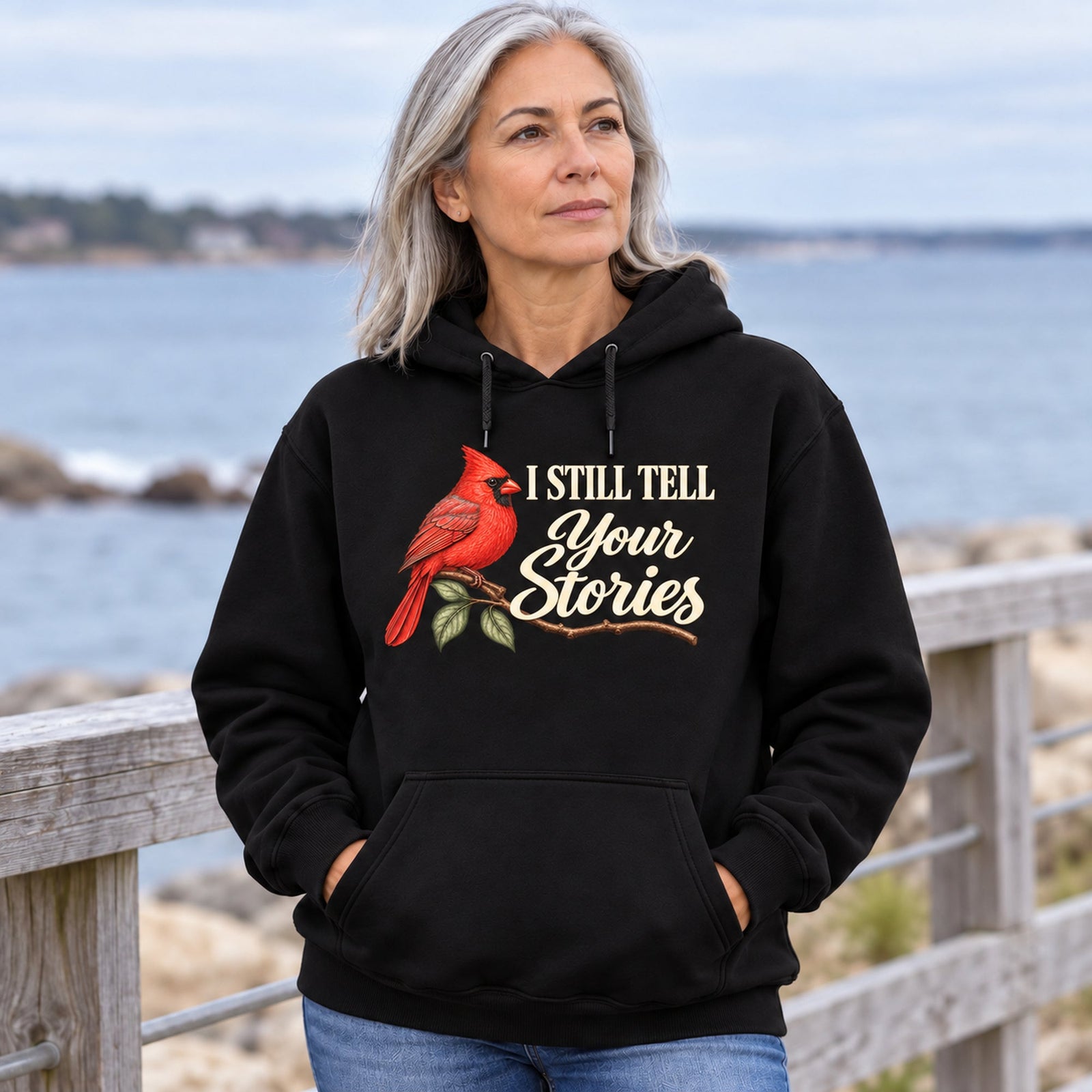 Reflective woman by the water wearing the revised I Still Tell Your Stories black hoodie with cardinal beside ivory lettering