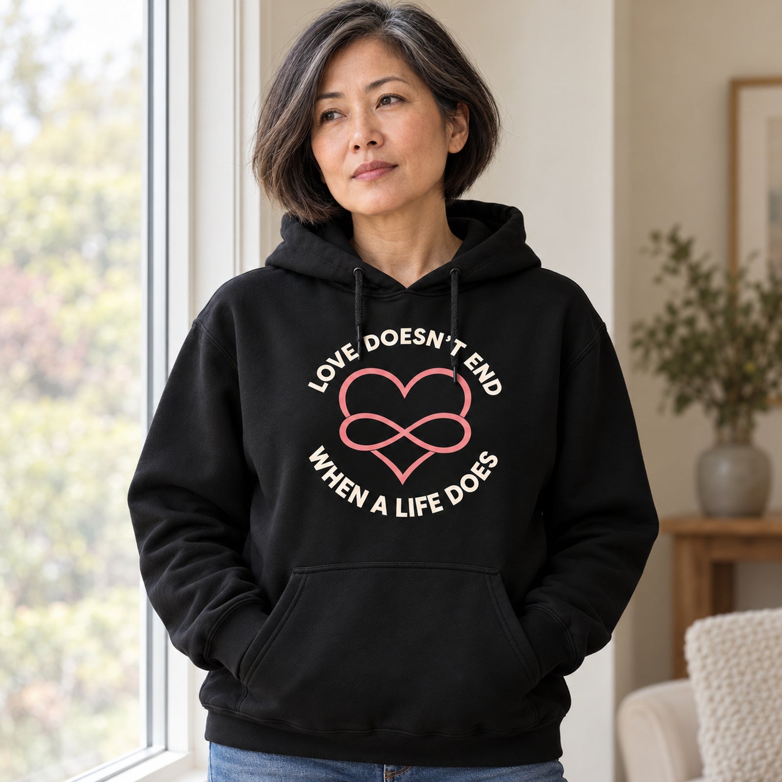 Reflective woman by a window wearing the black Love Doesn't End memorial hoodie